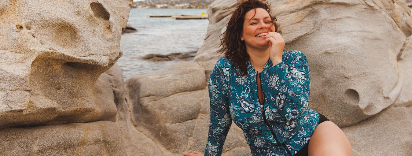 brunette woman poses by rocks wearing Sardinia teal and white floral long sleeve zip up rash vest