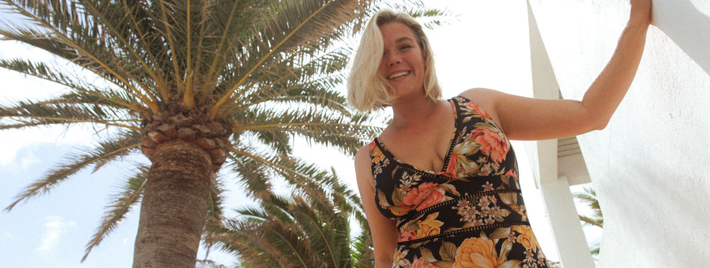 Woman with blonde hair poses by a palm tree wearing Frenchy Black floral V neck one piece swimsuit