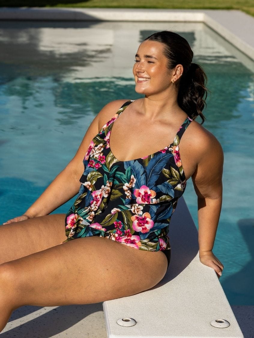 A woman wearing a floral Tiered tankini swimsuit sits on the edge of a swimming pool outdoors.