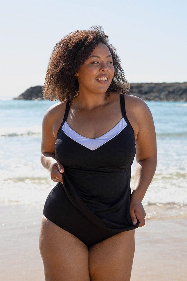 Brunette model stands on the beach in a black tankini. The tankini top is made of a textured fabric and has a white border along the bust line. 