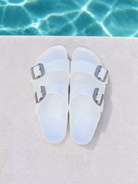 A pair of white double-strap slide sandals with silver buckles, displayed on a light beige surface with a swimming pool background visible at the top.