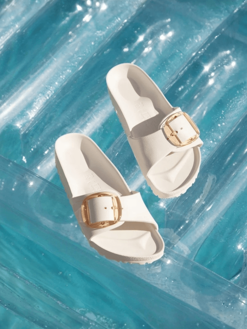 A pair of off-white slide sandals with gold-tone buckle details, arranged on a translucent blue inflatable pool float.