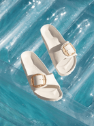A pair of off-white slide sandals with gold-tone buckle details, arranged on a translucent blue inflatable pool float.
