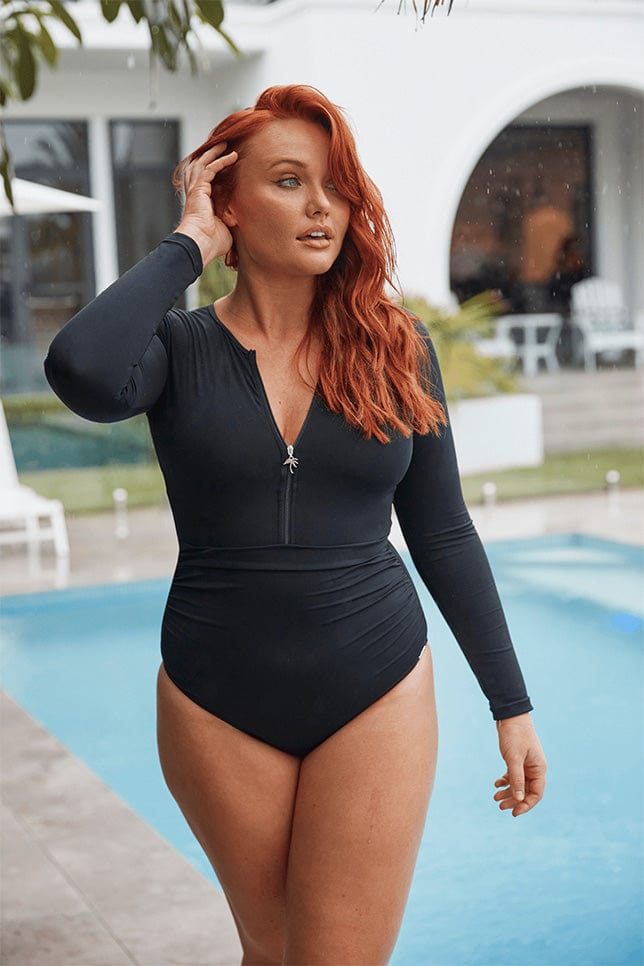 Red haired model stands poolside in a black chlorine resistant swimsuit with long sleeves and a zip front
