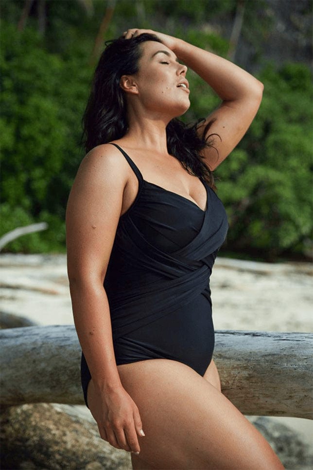 Model stands on beach in a black one piece. The swimsuit has fabric draping along the torso to create a waist defining and tummy flattering effect.