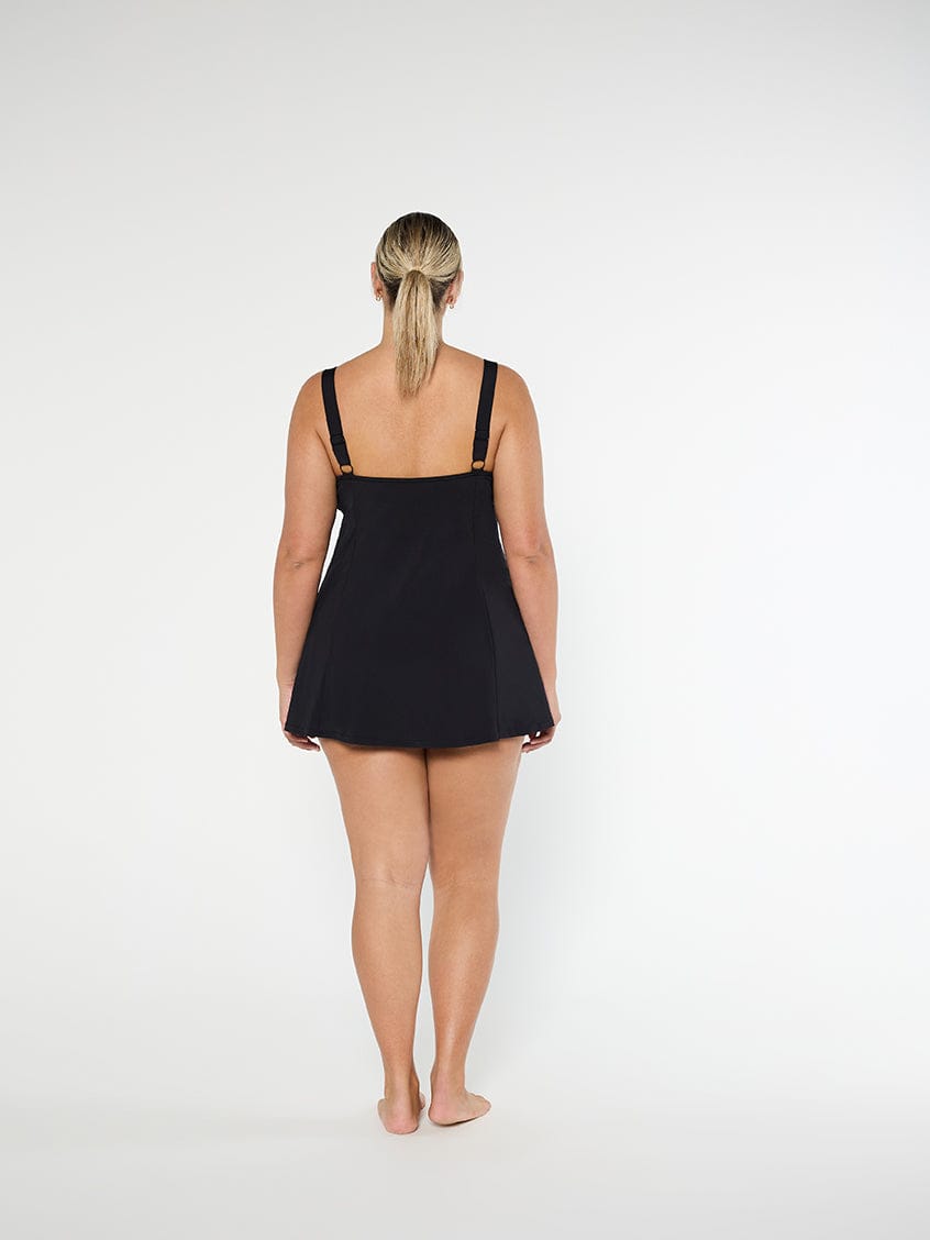 back of model wearing a black swim dress standing in front of a white wall