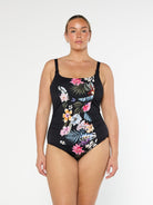 Blonde model shows the front of black floral one piece swimsuit with adjustable straps and supportive shelf bra