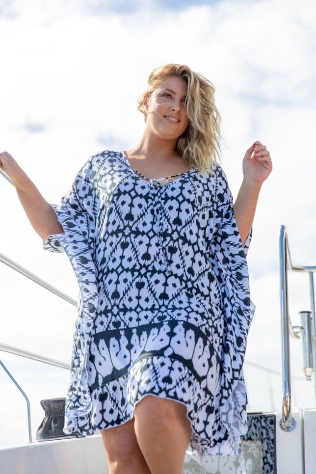Blonde woman stands on a boat in a white kaftan. The beach cover up has a black diamond tie dye like pattern