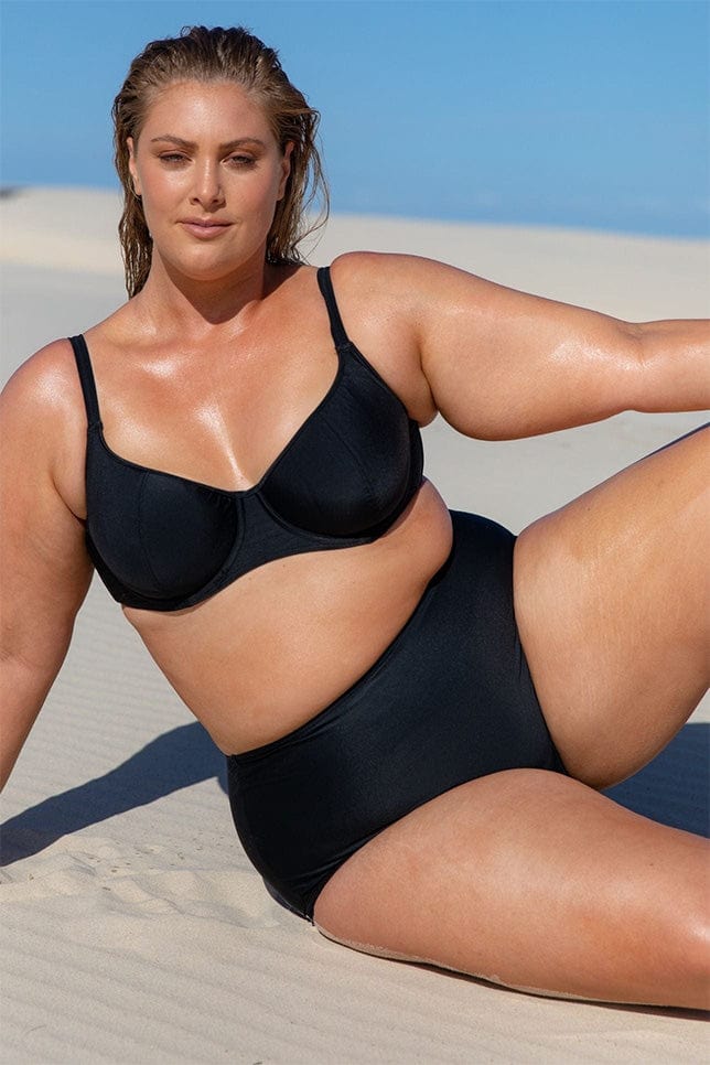 Woman poses on the sand. She wears a plain black bikini. The bikini bottoms are high waisted and full coverage, they provide excellent tummy control
