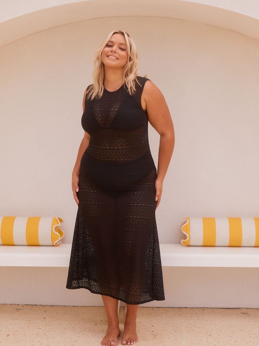 A woman modeling a black sleeveless crochet midi dress in front of a white arched wall with yellow and white striped pillows.