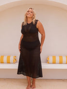 A woman modeling a black sleeveless crochet midi dress in front of a white arched wall with yellow and white striped pillows.