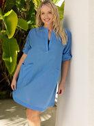 Woman wearing a blue dress standing next to a white column with greenery in the background