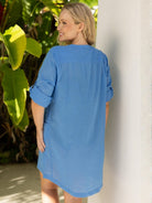 Woman wearing a blue dress leaning against a white column with greenery in the background