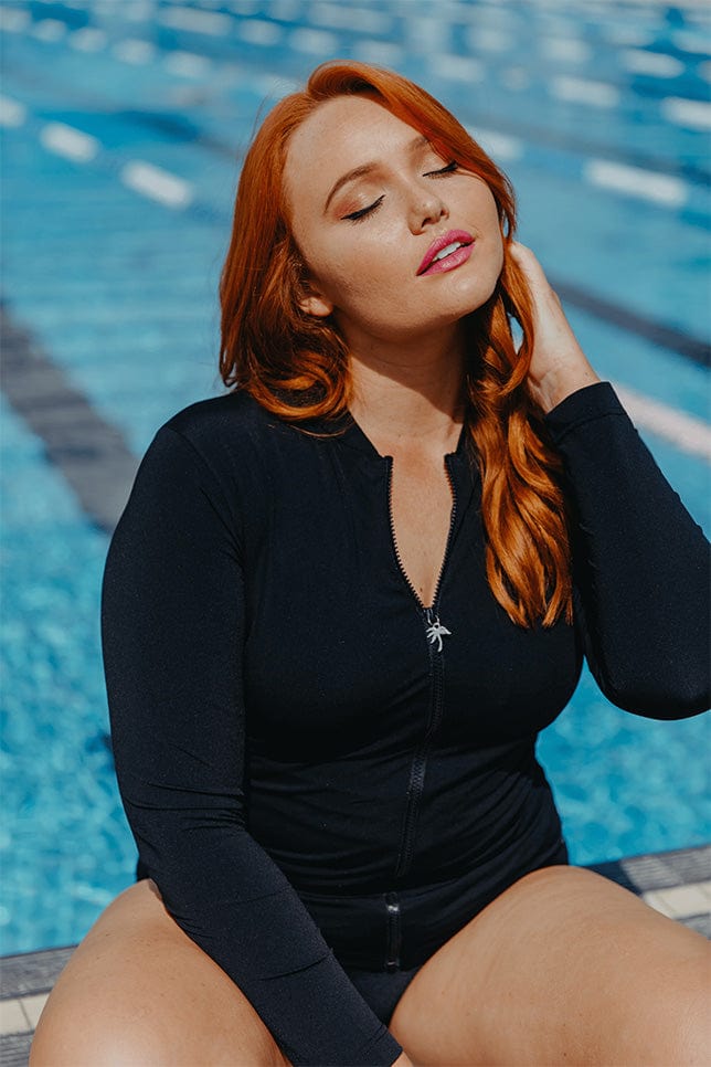 red haired model sits poolside with a black chlorine resistant rash vest on
