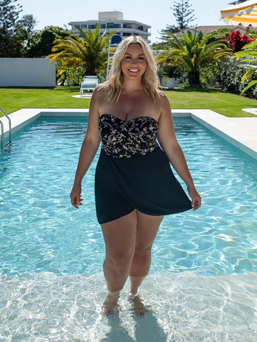 Woman standing by a pool wearing a strapless floral top and black skirt.