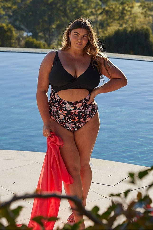Brunette woman stands poolside holding a pink sarong. She wears a Black one piece swimsuit. The swimsuit has a plain black top, floral printed bottom and a cut out  in the midriff. 