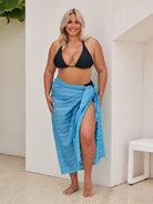 Woman wearing a black bikini top and blue sarong standing indoors.