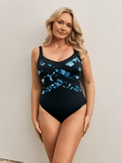 Blonde model standing by wall wearing a criss cross one piece swimsuit in blue and black floral