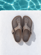 A pair of taupe-brown thong sandals with adjustable straps, laid flat on a white surface with a swimming pool background visible at the top.
