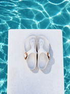 A pair of white slide sandals with metallic rose gold straps, placed on a white rectangular platform with a swimming pool background.