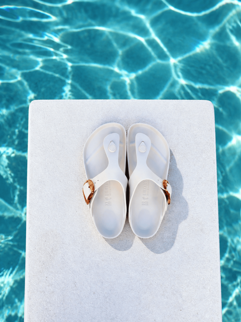 A pair of white slide sandals with metallic rose gold straps, placed on a white rectangular platform with a swimming pool background.