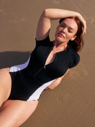Redhead model lays on the sand, wearing a high neck, cap sleeve swimsuit. The swimsuit, part of the Capriosca acapulco textured collection, has a zipped front and white panels on each side of the body.