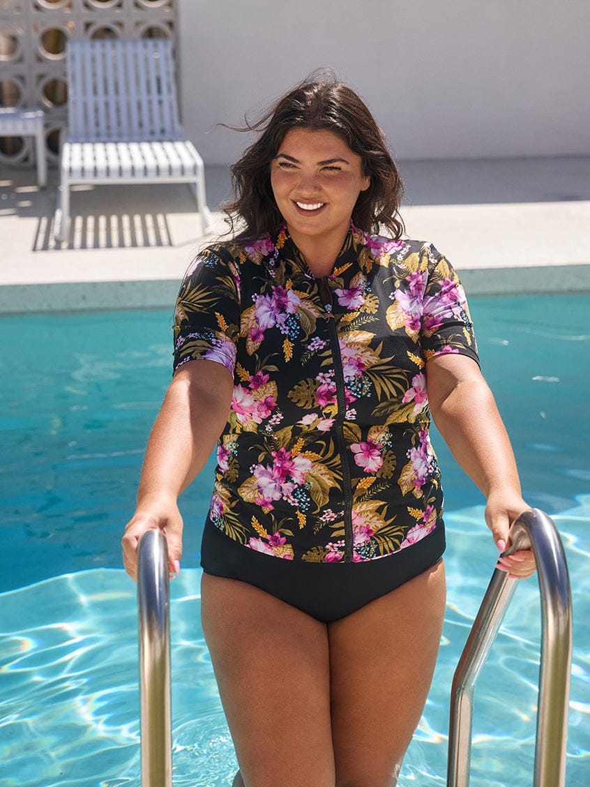 Woman in a floral shirt and black swimsuit standing by a pool.