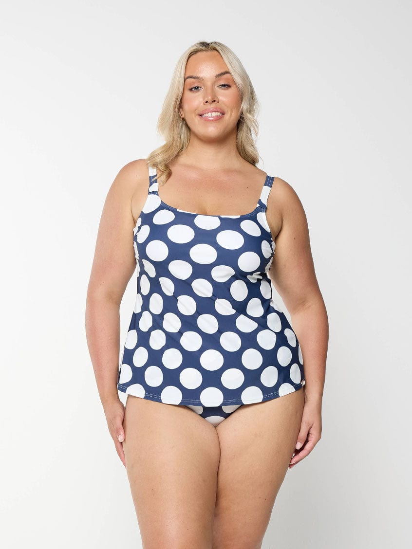 Blonde model shows the front of a flattering scoop neck tankini top with a navy and white polkadot pattern