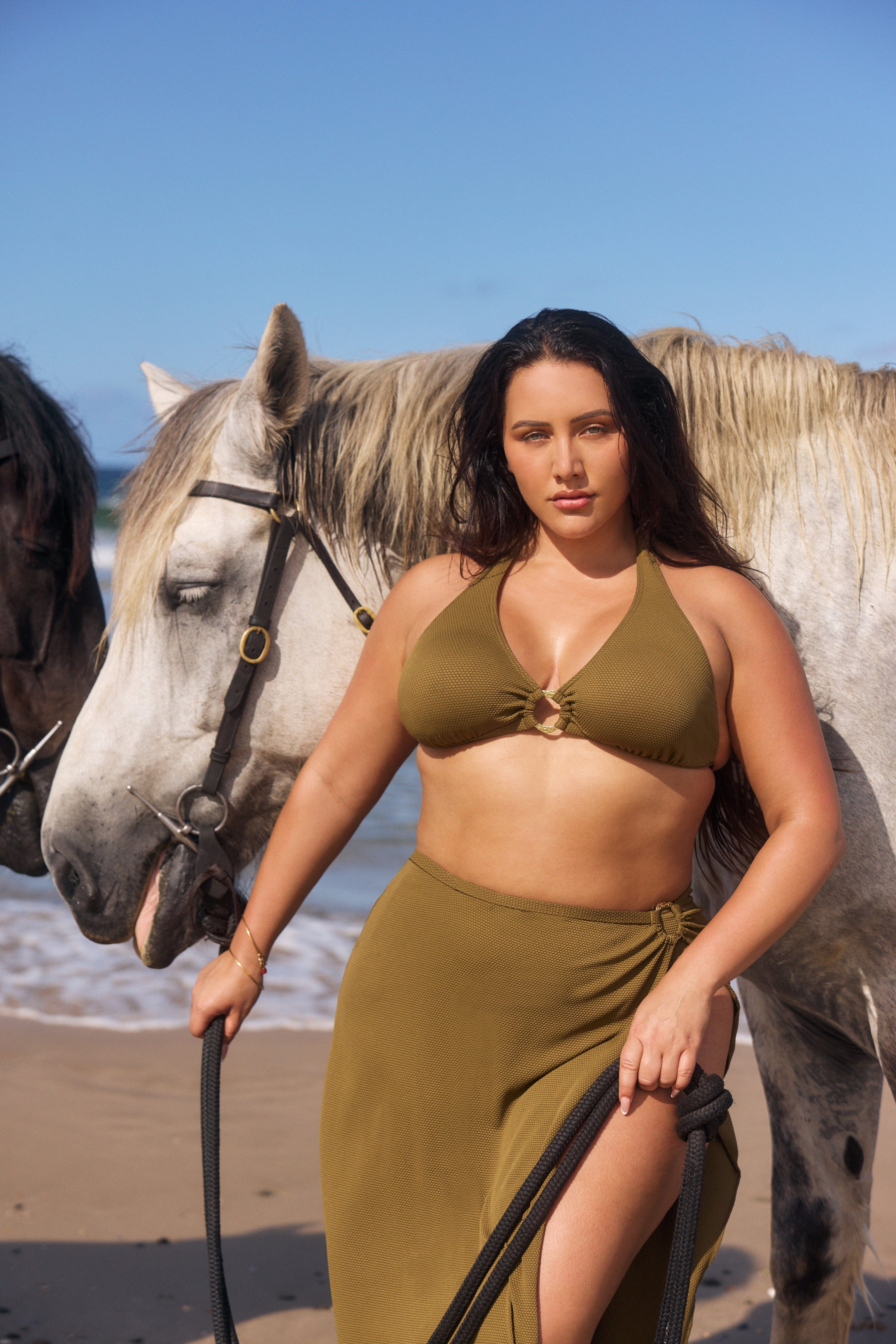 Woman in a green bikini and skirt standing on a beach with horses in the background