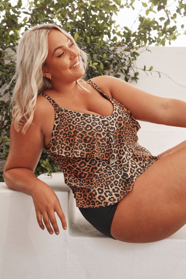 Blonde model sits on a white bench wearing a leopard print ruffled tankini. The tankini swimsuit has three frills down the front and is styled with a black bikini bottom.