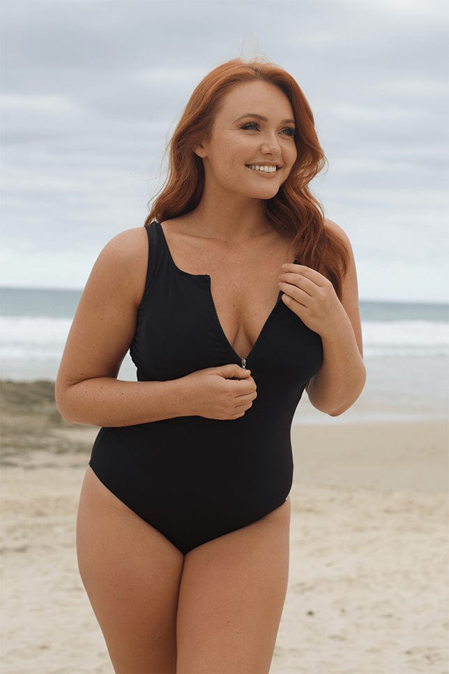 Model stands on beach in a plain black sleeveless one piece with a front zipper. She is holding the zip. The swimsuit looks good quality and is made of chlorine resistant fabric. 