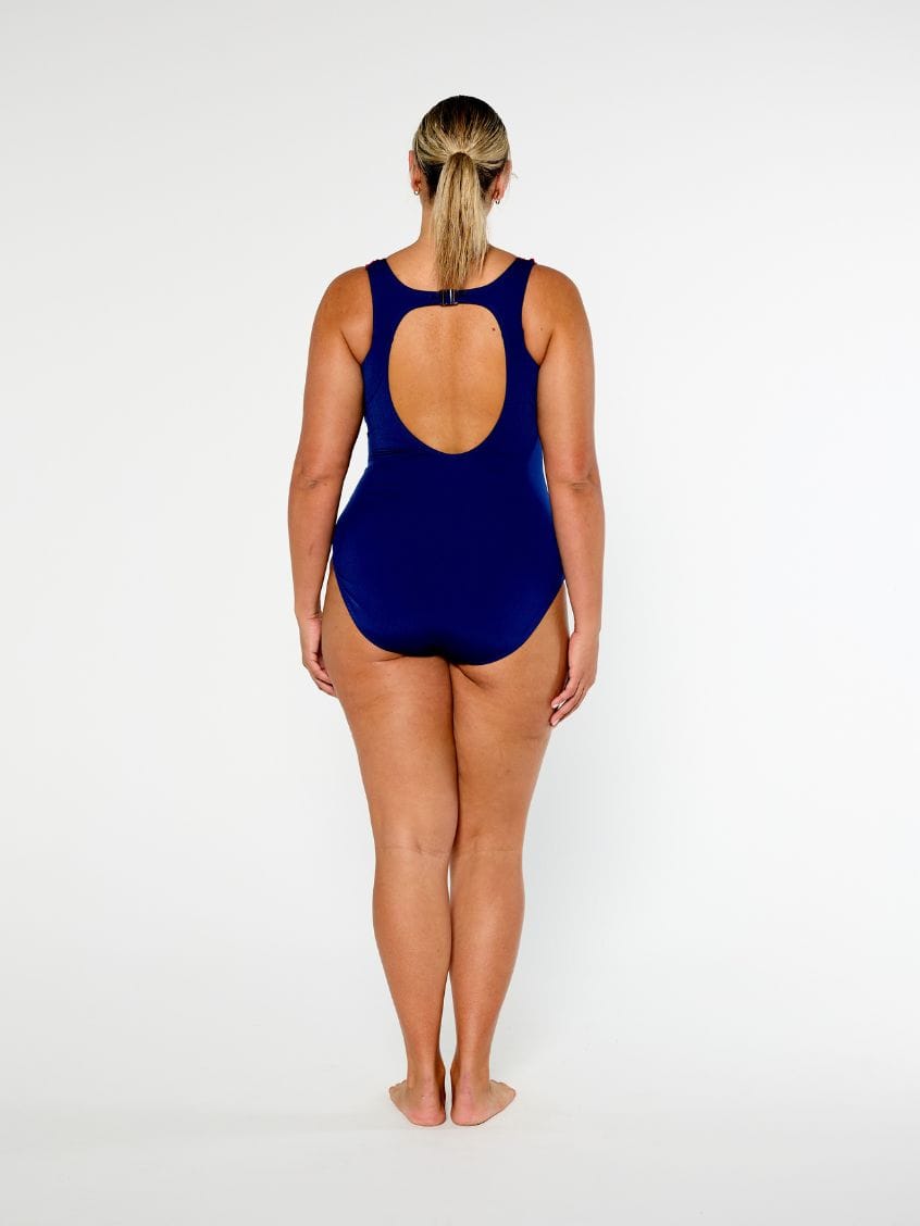 A woman standing with her back to the camera wearing a navy blue one-piece swimsuit with a large open back cutout, set against a plain white studio background.