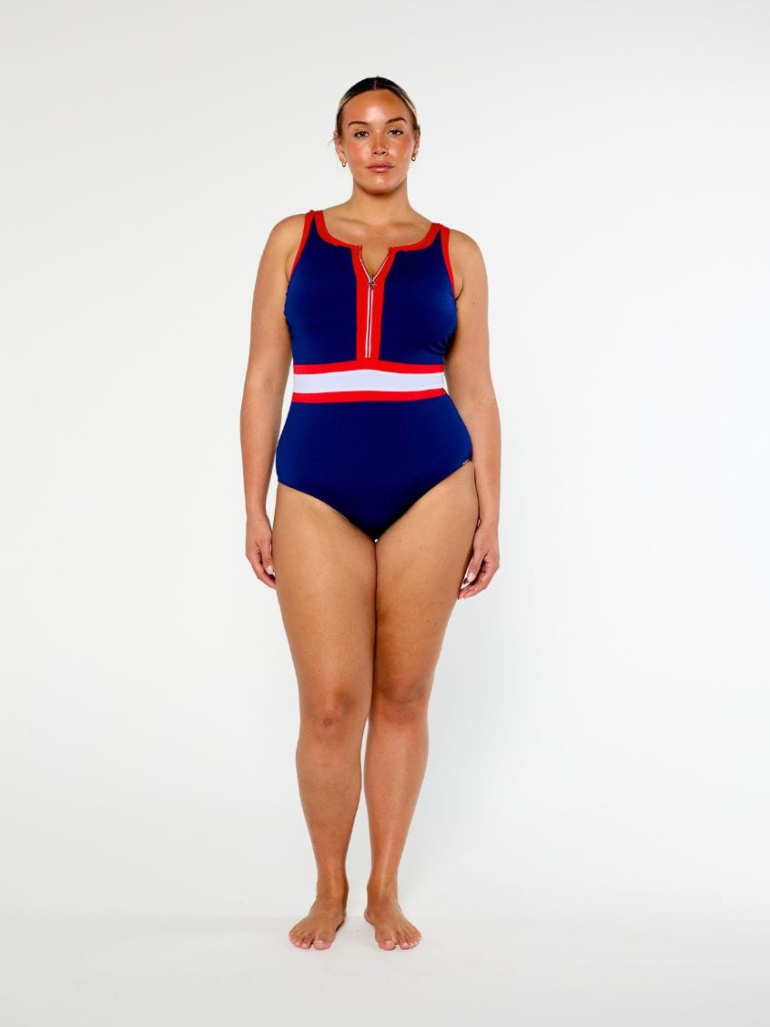 A woman modeling a one-piece swimsuit with a navy blue base and red and white striped accents on the neckline and waist, photographed against a plain light gray studio background.
