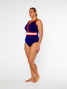 A woman modeling a one-piece swimsuit with a royal blue base and red and white stripe details, standing against a plain white studio background.