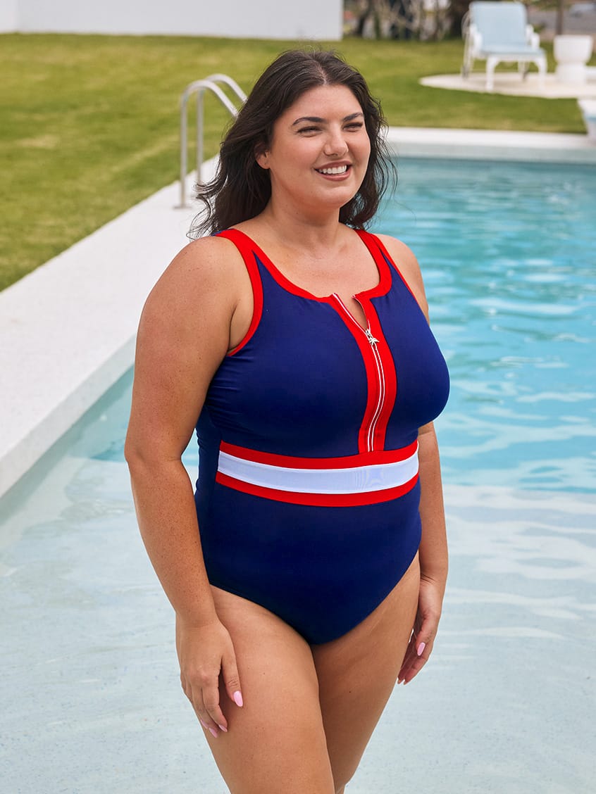 A woman wearing a navy blue one-piece swimsuit with red and white trim stands by a swimming pool. The swimsuit features a red and white zipper down the center chest, red trim along the neckline and armholes, and a horizontal red and white stripe across the waist.