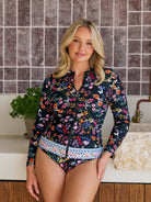 A woman modeling a long-sleeve floral swim zip-up top and matching bikini bottoms in a bathroom setting with a tiled wall and wooden counter.