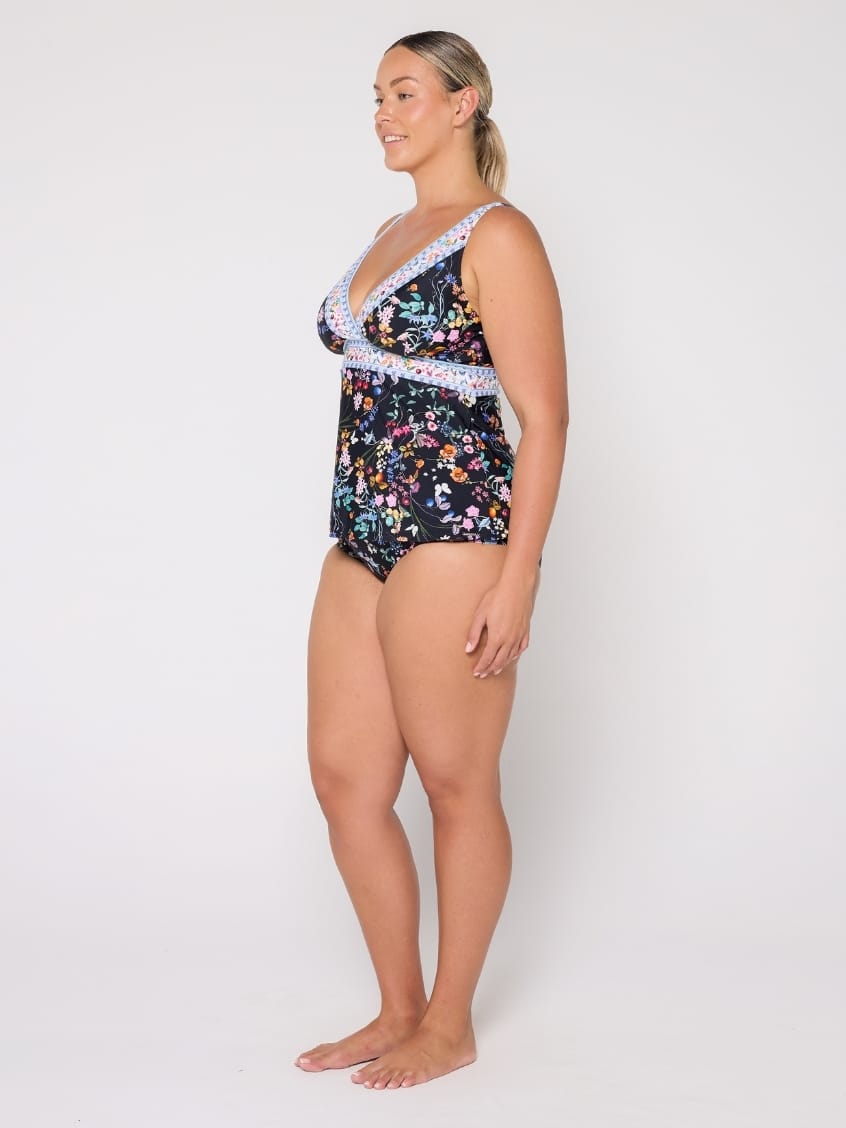A woman modeling a one-piece swimsuit with a dark navy floral print and light blue lace trim, standing against a plain white studio background.
