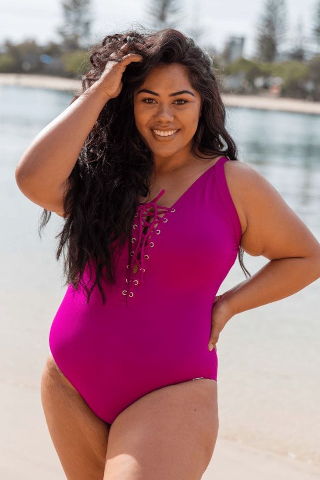 Model stands on beach, she wears a lightly textured magenta swimsuit. The one piece has a V neck cut out and lace up detail that is tied in a bow in the front.