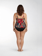 A woman is shown from behind wearing a one-piece swimsuit with a vibrant tropical floral print on the top and solid black bottoms, standing against a light gray studio background.