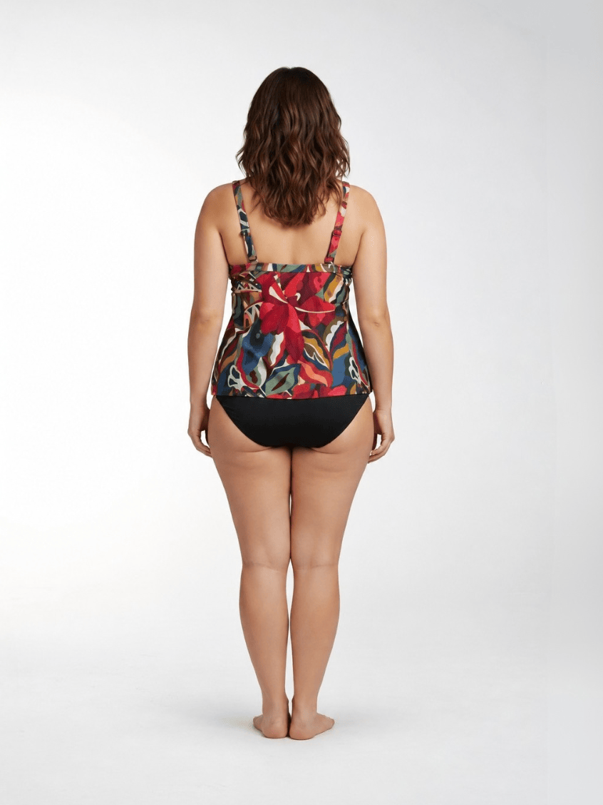 A woman is shown from behind wearing a one-piece swimsuit with a vibrant tropical floral print on the top and solid black bottoms, standing against a light gray studio background.
