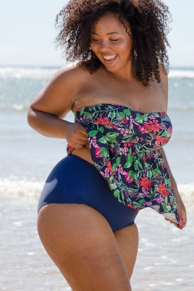 dark haired woman smiles on the beach while showing off her high waisted navy swim bottoms with a full coverage bottom.