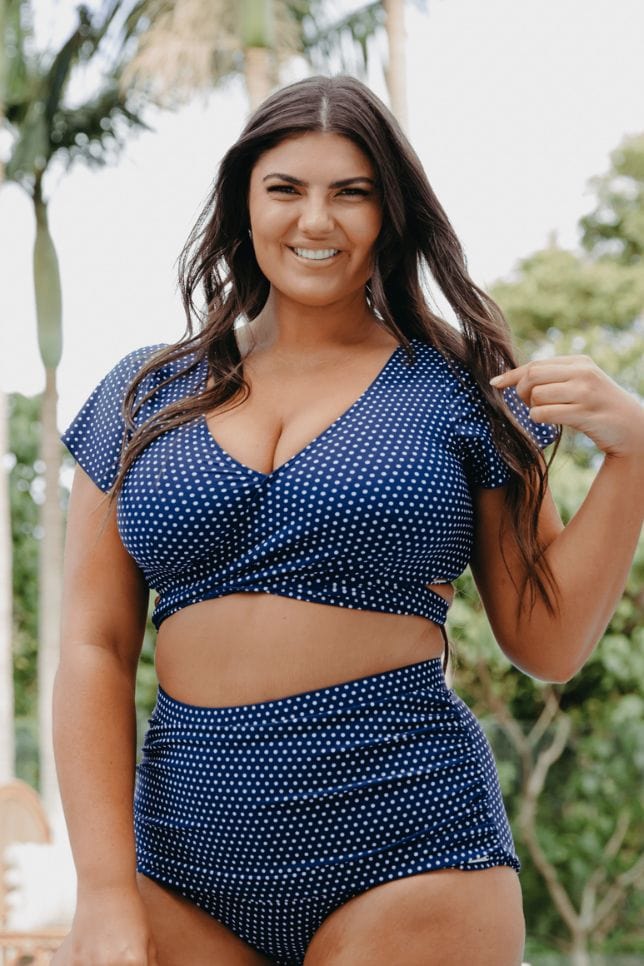 Model wears a navy swimsuit with white spots. The bikini top has short sleeves and a wrap feature at the front with a tie in the back.