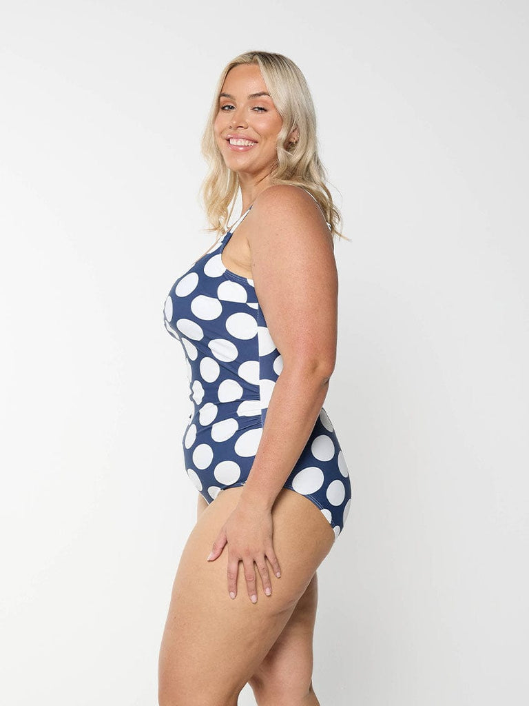 Blonde model shows the side of figure flattering one piece swimsuit in navy and white polkadot pattern