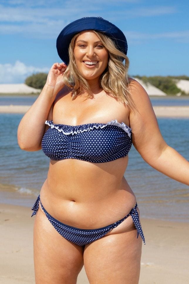 Blonde woman wearing a hat stands on the beach in a Navy and White Bikini. The bikini bottoms is low waisted and have ties on the side. The Bikini is navy with white spots.