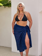 Woman wearing a black bikini top and navy blue sarong against a white wall.