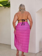 Woman wearing a pink sarong with pom poms and black bikini top against a white wall.