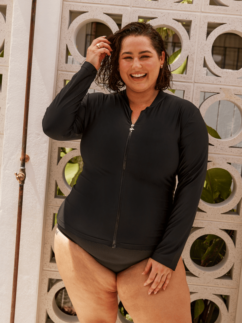 A woman wearing a black long-sleeve zip-up swim shirt and matching one-piece bottoms, standing in front of a decorative white wall with circular cutouts.