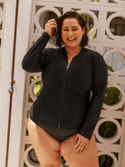 A woman wearing a black long-sleeve zip-up swim shirt and matching one-piece bottoms, standing in front of a decorative white wall with circular cutouts.