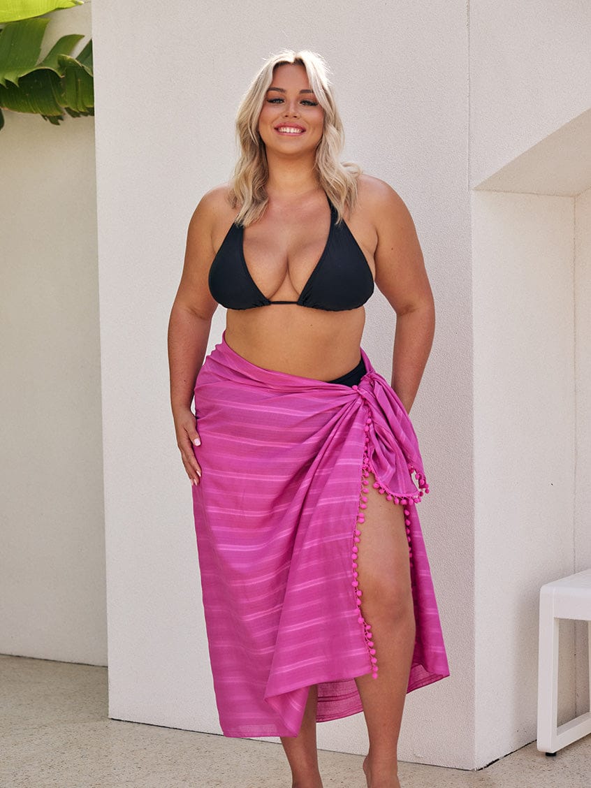 Woman wearing a black bikini top and pink striped sarong outdoors.