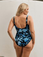 Back of model standing by wall wearing a scoop back tank one piece in floral print